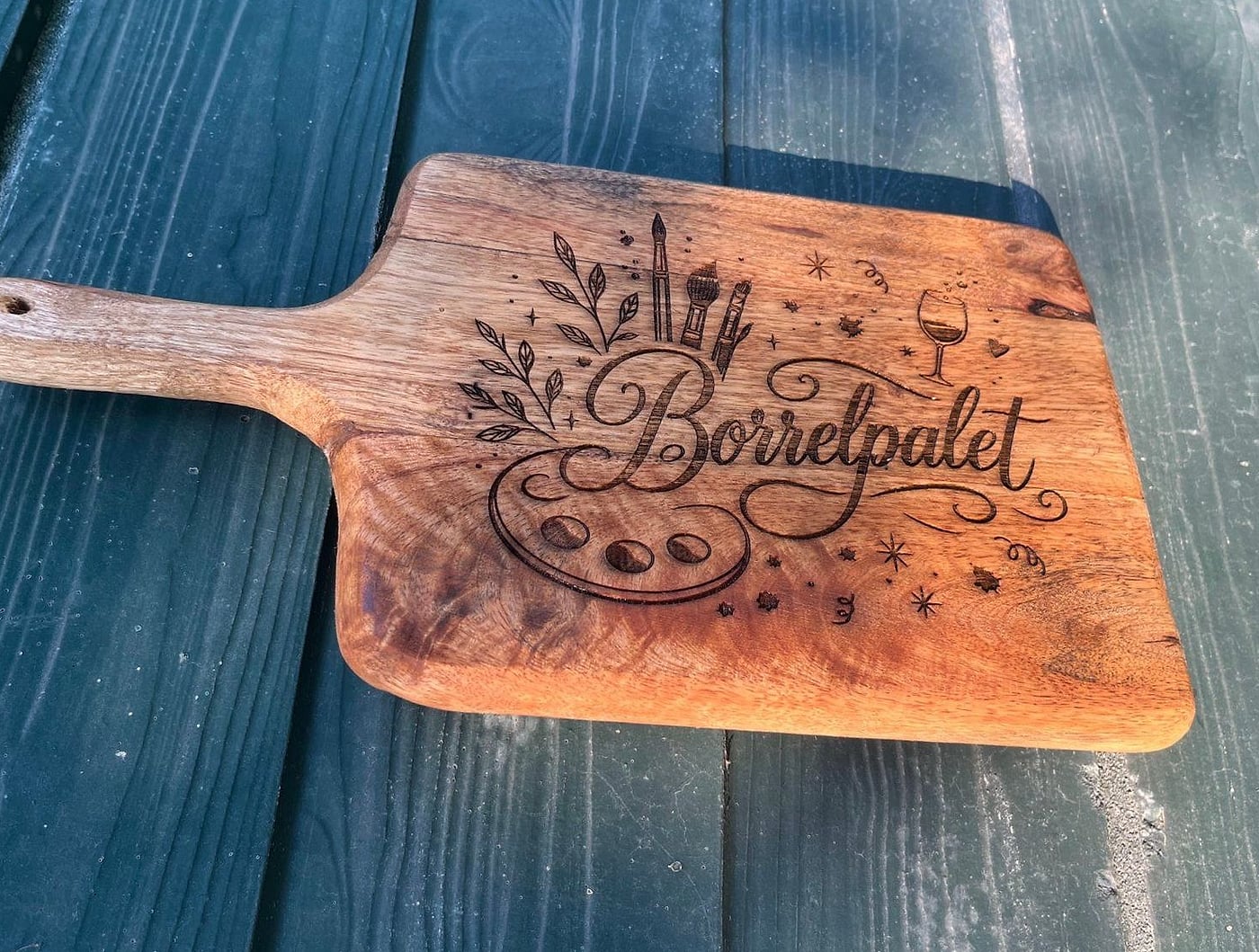 Houten borrelplank met handvat, gegraveerd met een schilderspalet, penselen, een wijnglas en de tekst Borrelpalet