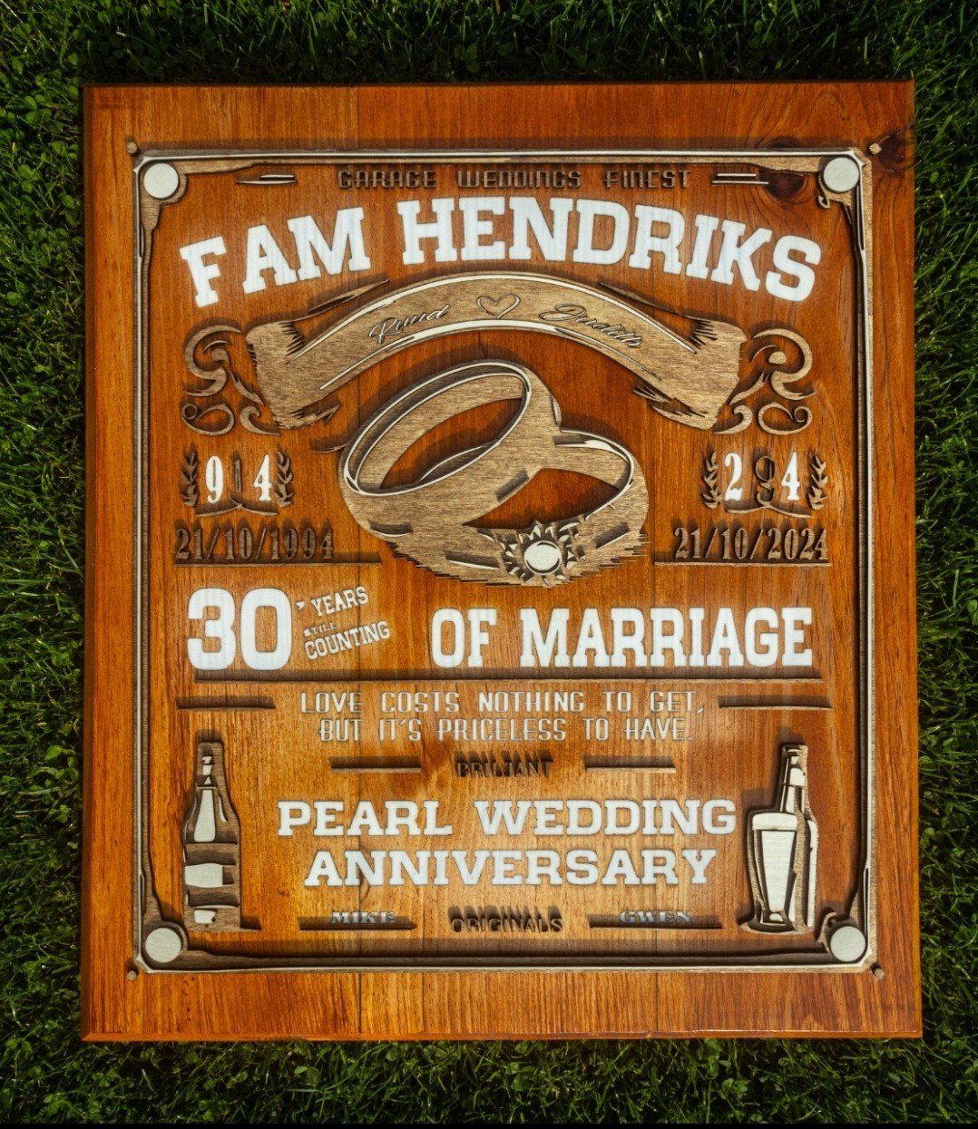 Gelaagd houten jubileumbord met trouwringen, data en tekst, in de zon op het gras