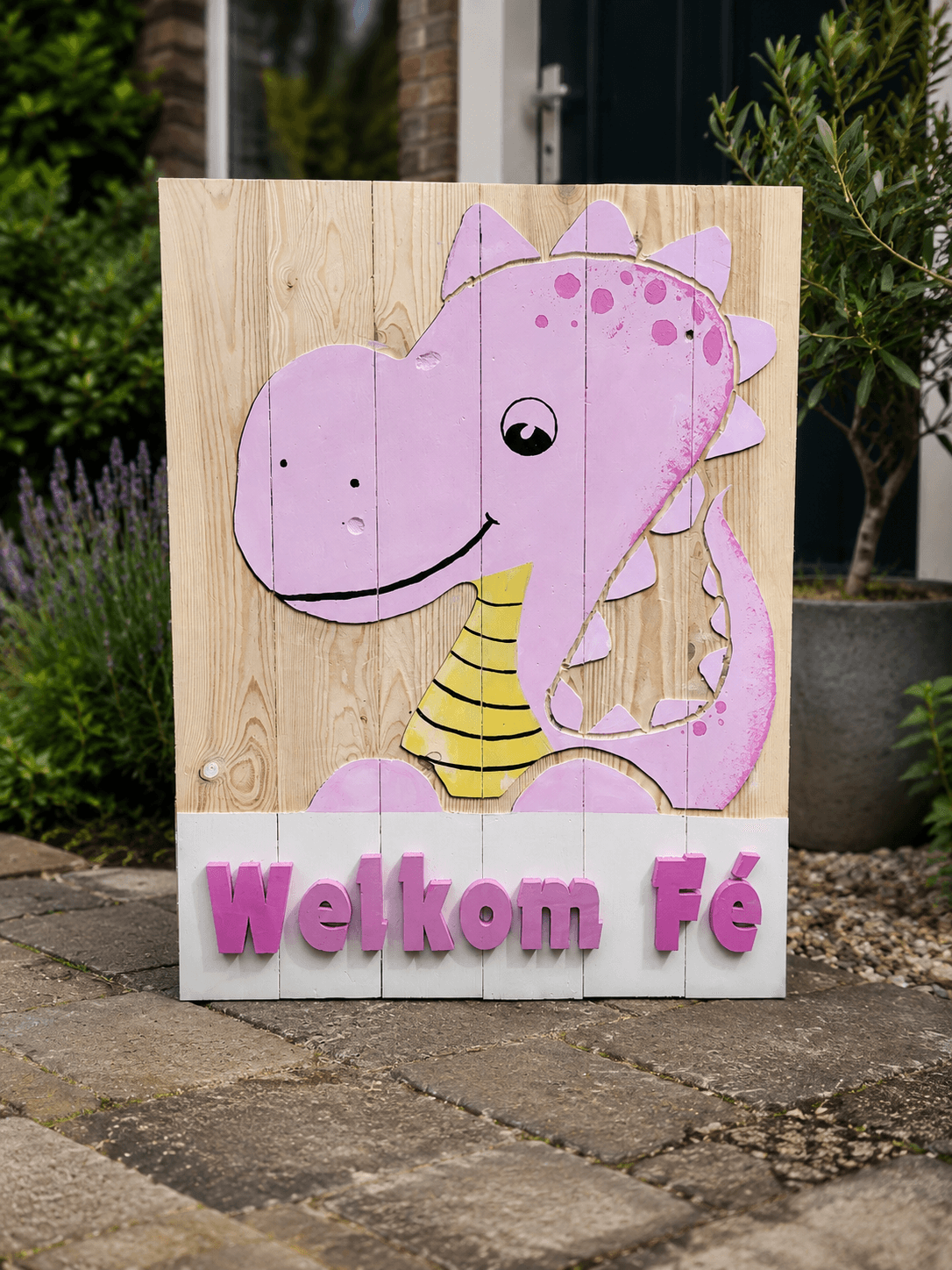 Houten bord met roze dinosaurus en gele hals, met tekst Welkom Fé, buiten in een tuin.